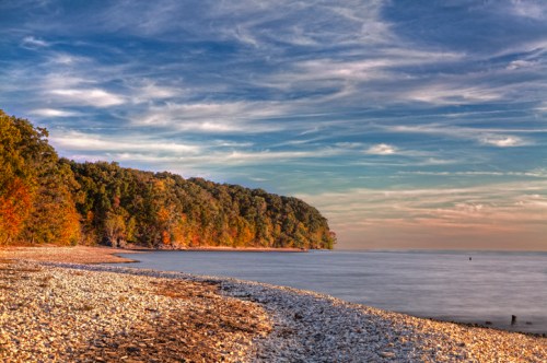 Serene beach scene with pebbles, colorful trees, and a calm water surface under a cloudy sky.