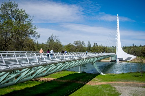 A modern suspension bridge spans a river, with people walking along it and greenery surrounding the area.