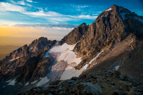 A stunning mountain landscape at sunset, featuring rocky peaks and patches of snow under a colorful sky.