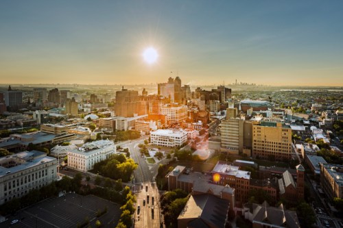 Aerial view of a city skyline at sunset, with buildings and roads illuminated by warm sunlight.