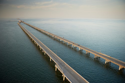 Aerial view of a long pier extending into calm waters, with a road running alongside it under a clear sky.