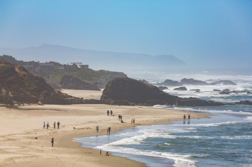 A scenic beach with people walking along the shore, rocky formations, and gentle waves under a clear blue sky.
