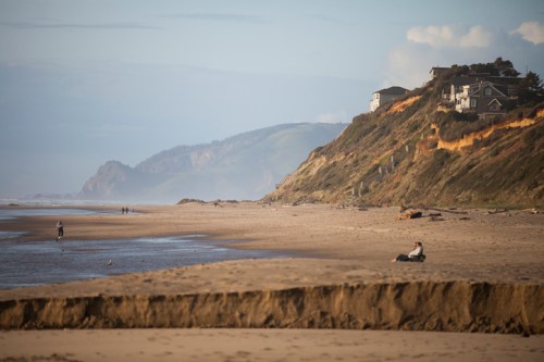 A sandy beach with a cliffside and houses in the background, people walking along the shore under a clear sky.