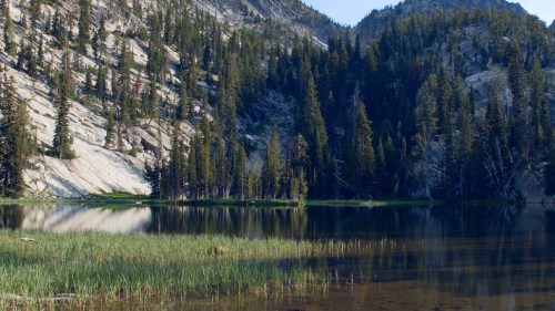 A serene lake surrounded by tall trees and rocky mountains, reflecting the landscape in calm waters.