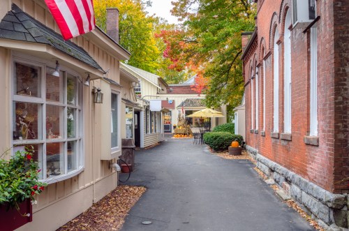 Charming alleyway lined with shops, colorful autumn leaves, and an American flag, creating a cozy fall atmosphere.