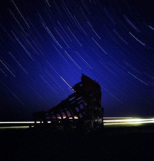 A shipwreck silhouette against a starry night sky, with star trails and faint light streaks in the background.