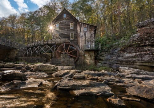 A historic wooden mill with a water wheel, surrounded by trees and rocks, illuminated by sunlight in a serene landscape.