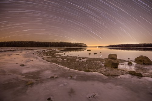 A serene lake at night with star trails above, surrounded by rocky shores and a hint of ice along the water's edge.