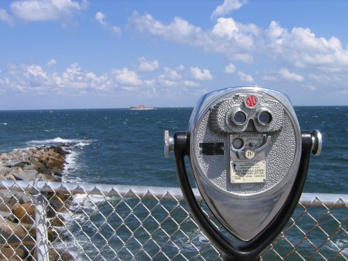 A coin-operated binocular viewer overlooking a rocky shoreline and distant island under a blue sky with clouds.