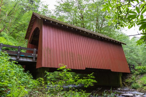 A red covered bridge stands over a stream, surrounded by lush green trees and foliage.