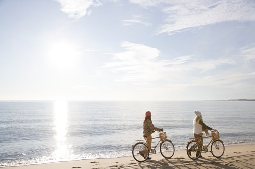Two people ride bicycles along a serene beach at sunset, with calm waters and a clear sky in the background.