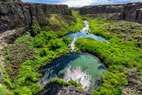 A lush green canyon with a winding river, surrounded by rocky cliffs under a partly cloudy sky.