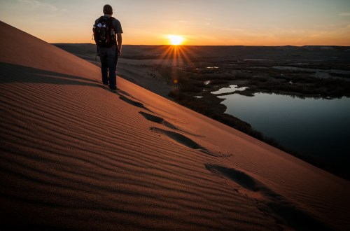 A person walks along a sand dune at sunset, with footprints leading to a serene river below.