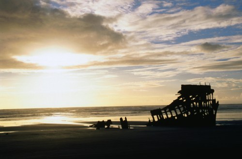 Silhouette of a shipwreck on a beach at sunset, with dramatic clouds and calm waters in the background.