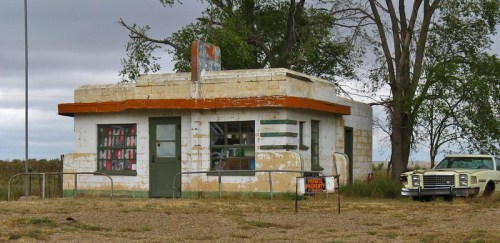 Abandoned building with peeling paint, surrounded by trees and an old car parked nearby. Overcast sky in the background.