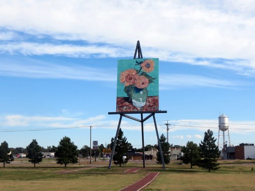 A large outdoor painting of sunflowers on a sign, set against a blue sky and green grass with trees nearby.