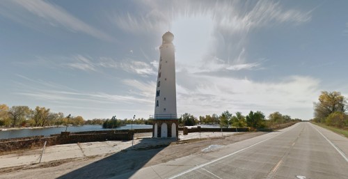 A tall white lighthouse stands beside a calm body of water, with a road running alongside it under a bright sky.