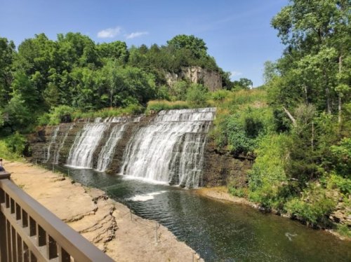 A serene waterfall cascading over rocky ledges, surrounded by lush greenery and a clear blue sky.