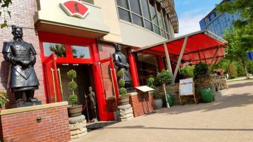 Exterior of a restaurant featuring red doors, large statues, and potted plants, set against a clear blue sky.