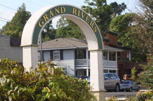 A sign reading "Grand Rivers" arches over a street scene with houses and greenery in the background.