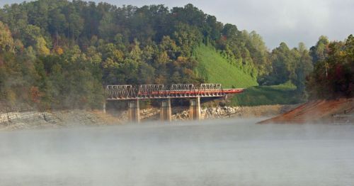 A misty lake scene with a bridge over water, surrounded by lush green hills and trees in the background.