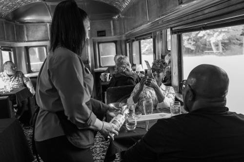 A waitress serves drinks to two diners in a vintage train car, with other patrons enjoying their meals in the background.