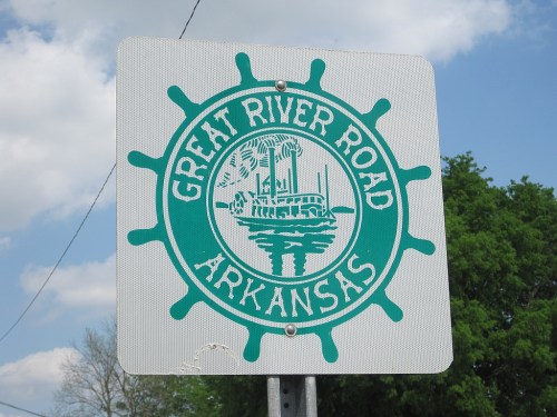 Sign for Great River Road in Arkansas, featuring a steamboat design and green color against a cloudy sky.