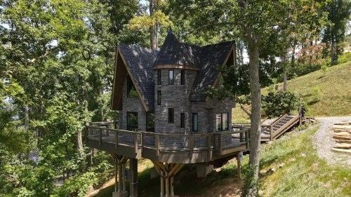 A unique stone house on stilts surrounded by trees, featuring a sloped roof and large windows.