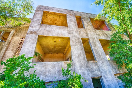Abandoned building with large empty windows, surrounded by greenery under a clear blue sky.