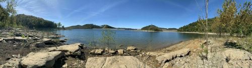 A panoramic view of a calm lake surrounded by trees and rocky shores under a clear blue sky.