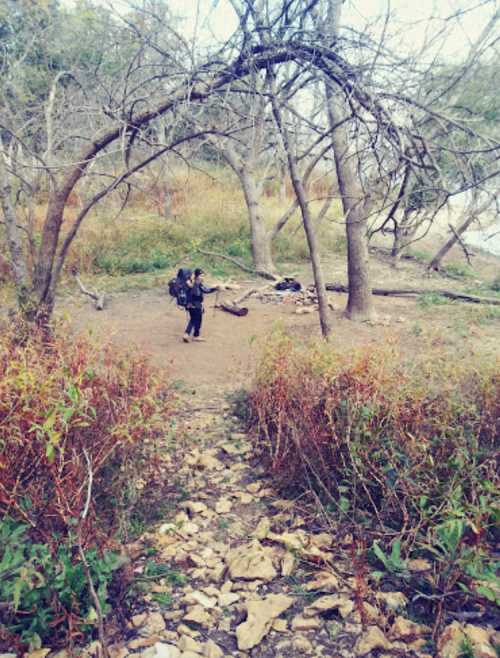 A hiker with a backpack walks along a rocky path surrounded by trees and colorful foliage.