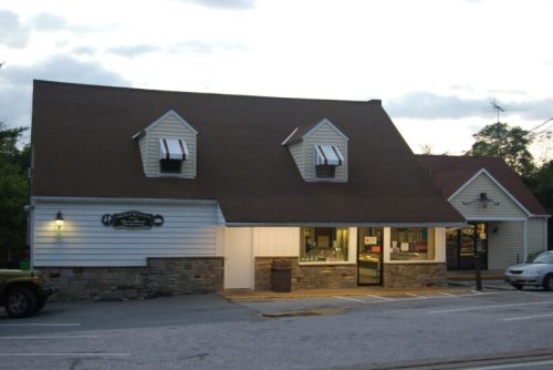 A quaint, two-story building with a sloped roof, featuring a stone facade and large windows, set in a parking area.