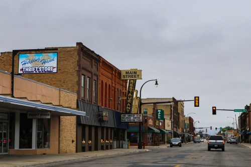 A street view of a small town featuring shops, a thrift store, and a restaurant sign on a cloudy day.