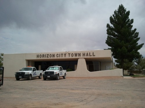 Horizon City Town Hall building with two parked trucks in front, under a cloudy sky.