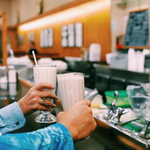 Two hands hold tall glasses of milkshakes at a diner counter, with a blurred background of the restaurant.