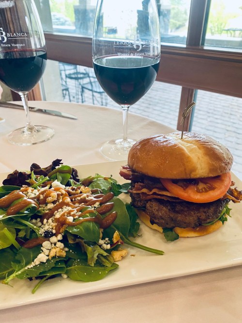 A gourmet burger with bacon, tomato, and cheese, served with a side salad and two glasses of red wine.