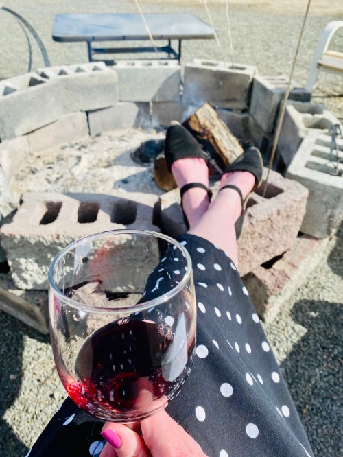 A person relaxes by a fire pit, holding a glass of red wine, with their feet resting on the edge of the pit.