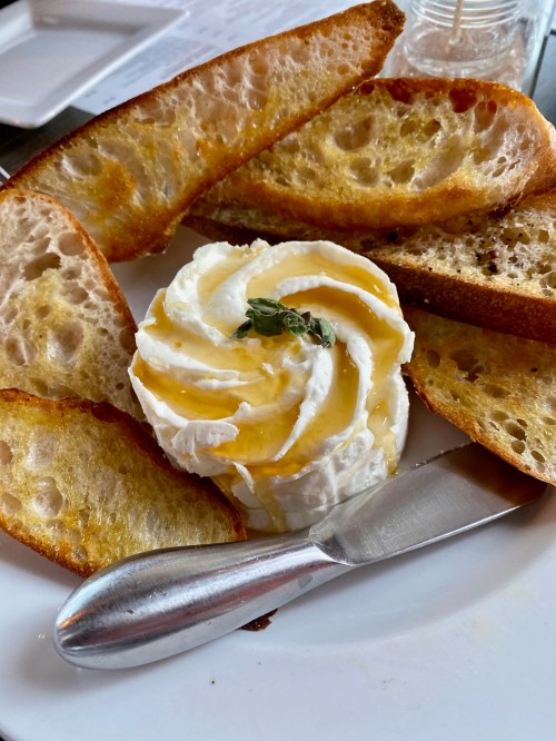 A plate with slices of toasted bread and a swirl of cream topped with honey and a sprig of herb, accompanied by a knife.