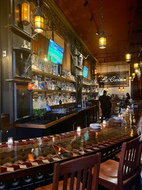 A cozy bar with a wooden counter, shelves of liquor, and TVs showing a sports game, illuminated by warm pendant lights.