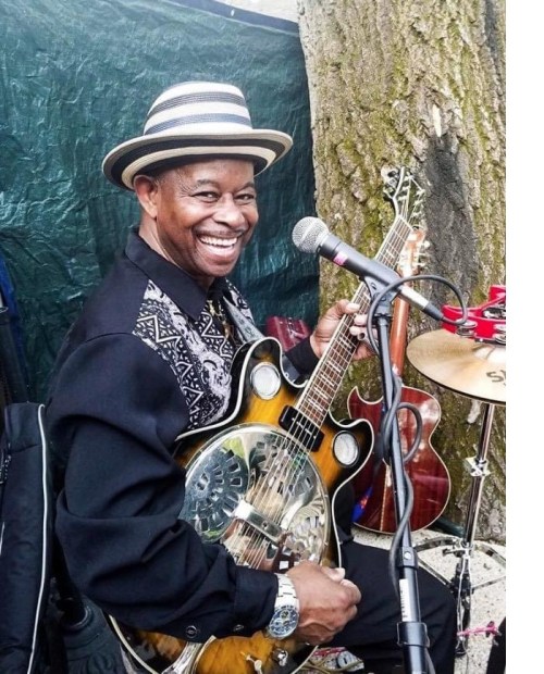 A smiling musician in a hat plays a resonator guitar, with a microphone and drums in the background.