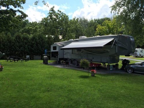A parked RV with an awning in a grassy area, surrounded by trees and outdoor furniture.