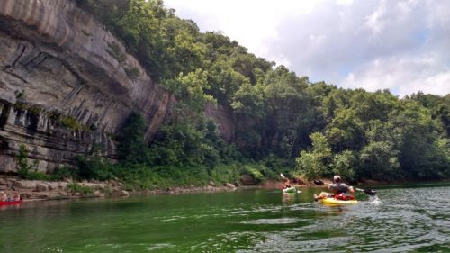 Two kayakers paddle on a calm river surrounded by lush green trees and steep rocky cliffs under a partly cloudy sky.