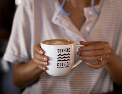 A person holding a white coffee mug with "SURFERS COFFEE" logo, featuring latte art on top.