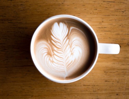 A close-up of a latte in a white cup, featuring intricate leaf-shaped foam art on a wooden table.