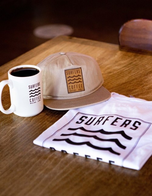 A coffee mug, cap, and t-shirt with a "Surfers Coffee" logo on a wooden table.