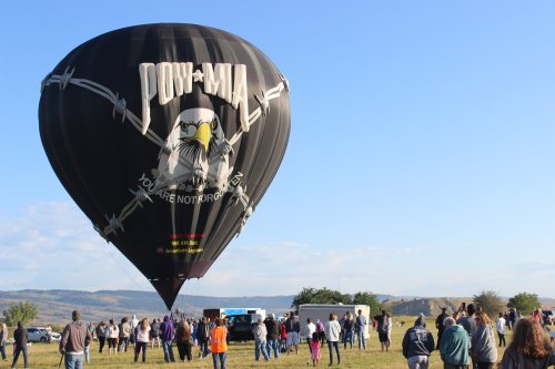 A black hot air balloon with an eagle design rises above a crowd on a clear day, with mountains in the background.