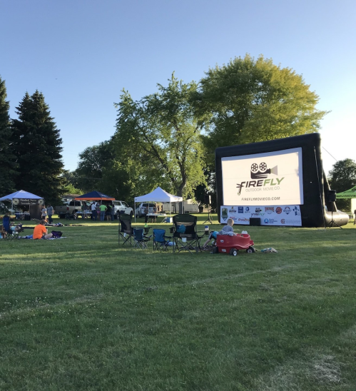 Outdoor event with a large screen displaying "FIREFLY," surrounded by tents and people sitting on the grass.