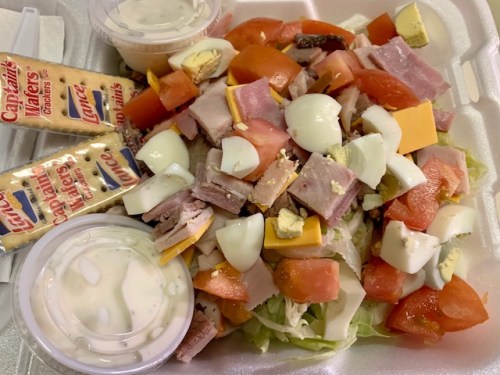 A salad with chopped ham, cheese, tomatoes, hard-boiled eggs, and two cups of dressing, served with crackers.