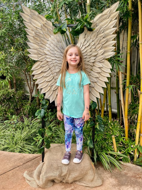 A smiling girl stands in front of large decorative wings, surrounded by greenery and bamboo. She wears a light blue shirt and colorful leggings.
