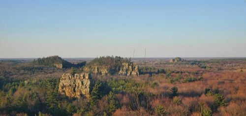 A panoramic view of rocky formations surrounded by trees and open fields under a clear blue sky.